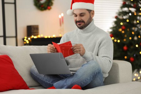 Weihnachten online feiern und Geschenke per Post austauschen. Mann mit Weihnachtsmütze liest Grußkarte während Videoanruf auf Laptop zu Hause