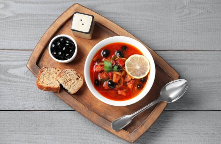 Fleischsoljanka-Suppe mit Würstchen, Oliven und Gemüse in Schüssel serviert auf grauem Holztisch, Draufsicht