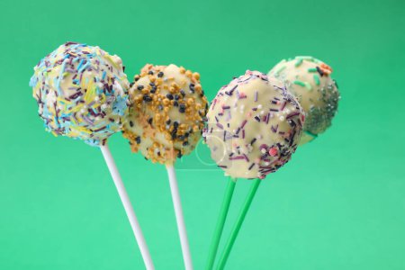 Sweet cake pops decorated with sprinkles on green background, closeup. Delicious confectionery