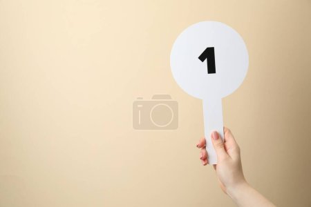 Woman holding auction paddle with number 1 on beige background, closeup. Space for text