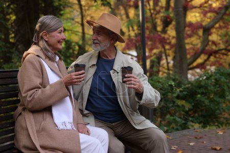 Téléchargez les photos : Couple sénior affectueux avec des tasses de café passer du temps ensemble sur le banc dans le parc d'automne - en image libre de droit