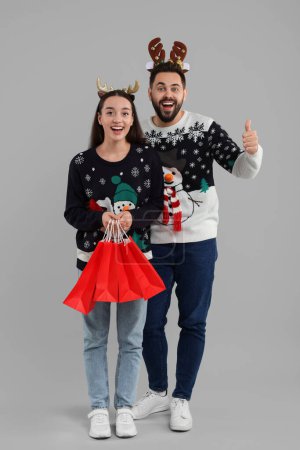 Glückliches junges Paar in Weihnachtspullis und Rentier-Stirnbändern mit Einkaufstaschen auf grauem Hintergrund