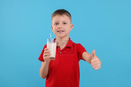 Téléchargez les photos : Mignon garçon avec verre de lait frais montrant pouce vers le haut sur fond bleu clair - en image libre de droit