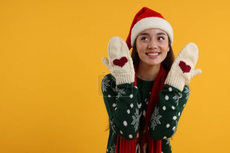 Happy young woman in Christmas sweater, Santa hat and knitted mittens on orange background. Space for text