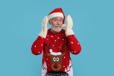 Senior man in Christmas sweater, Santa hat and knitted mittens on light blue background
