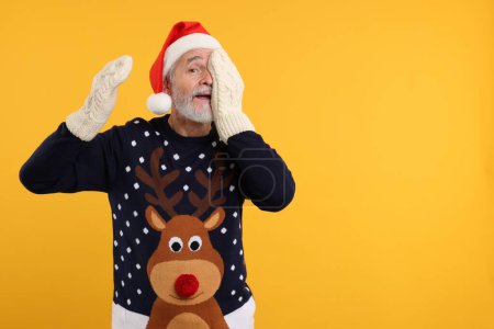 Senior man in Christmas sweater, Santa hat and knitted mittens on orange background. Space for text