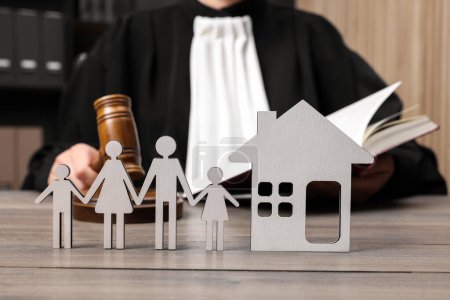 Family law. Judge with gavel and book sitting at wooden table, focus on figures of parents, children and house