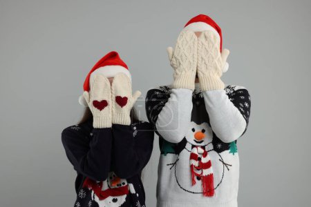 Young couple in Christmas sweaters and Santa hats covering faces with hands in knitted mittens on grey background
