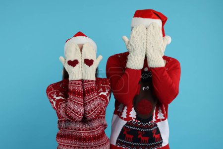 Young couple in Christmas sweaters and Santa hats covering faces with hands in knitted mittens on light blue background