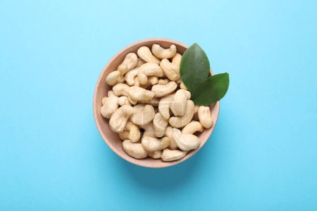 Tasty cashew nuts and green leaves in bowl on light blue background, top view