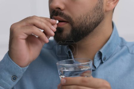 Man with glass of water taking antidepressant pill on light grey background, closeup