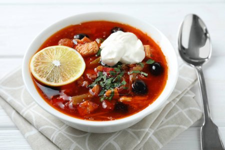 Meat solyanka soup with sausages, olives, vegetables and spoon on white wooden table, closeup