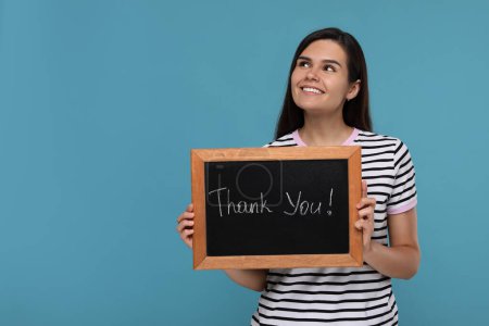Happy woman holding small chalkboard with phrase Thank You on light blue background, space for text