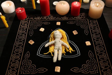 Muñeca vudú perforada con alfileres, runas y velas en alfombra negra. Ceremonia de maldición