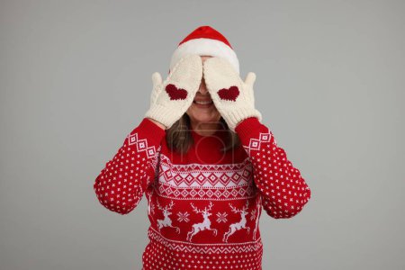 Heureuse femme âgée en pull de Noël et chapeau de Père Noël couvrant son visage avec les mains en moufles tricotées sur fond gris