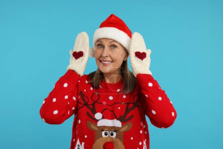 Femme âgée en pull de Noël, chapeau de Père Noël et mitaines tricotées sur fond bleu clair