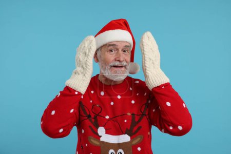 Hombre mayor en jersey de Navidad, sombrero de Santa y mitones de punto sobre fondo azul claro
