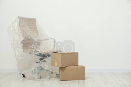 Téléchargez les photos : Chaise de bureau moderne recouverte de film plastique et de boîtes près du mur blanc à l'intérieur. Espace pour le texte - en image libre de droit