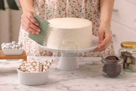 Woman using scraper to decorate cake at white marble table in kitchen, closeup