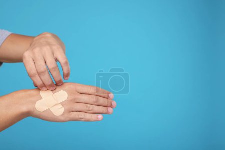 Man putting sticking plasters onto hand on light blue background, closeup. Space for text