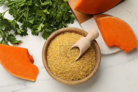Raw bulgur in bowl, scoop, pieces of pumpkin and parsley on white table, flat lay