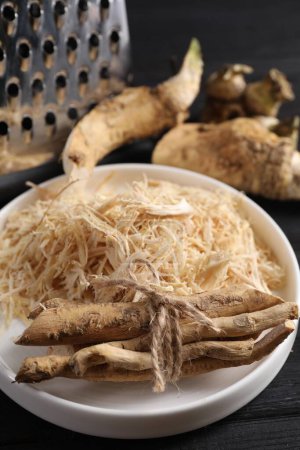 Plate with whole and grated horseradish roots on black wooden table, closeup