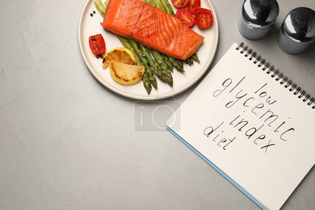 Cuaderno con palabras Low Glycemic Index Dieta y plato de sabrosa mesa gris salmón a la parrilla, puesta plana. Espacio para texto
