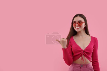 Pink look. Beautiful woman in heart shaped sunglasses pointing at something on color background, space for text