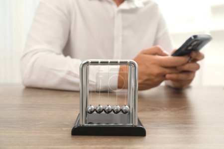 Photo for Newton's cradle and man using smartphone on wooden table, selective focus. Physics law of energy conservation - Royalty Free Image