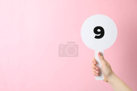 Woman holding auction paddle with number 9 on pink background, closeup. Space for text