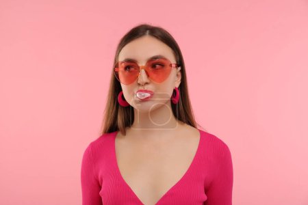 Pink look. Beautiful woman in heart shaped sunglasses blowing bubble gum on color background