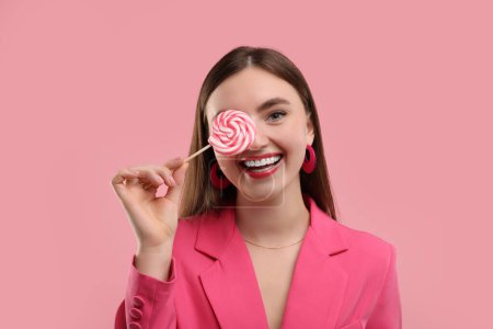 Beautiful woman in pink clothes covering eye with lollipop on color background
