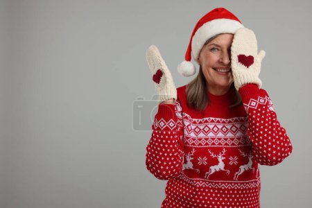 Mujer mayor feliz en jersey de Navidad, sombrero de Santa y mitones de punto sobre fondo gris. Espacio para texto