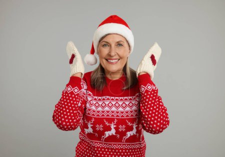 Joyeux senior femme en pull de Noël, chapeau de Père Noël et mitaines tricotées sur fond gris