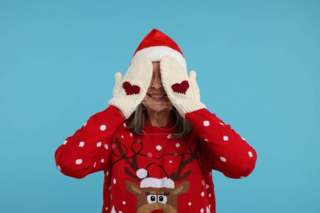 Mujer mayor en jersey de Navidad y la cara de la cubierta del sombrero de Santa con las manos en mitones de punto sobre fondo azul claro