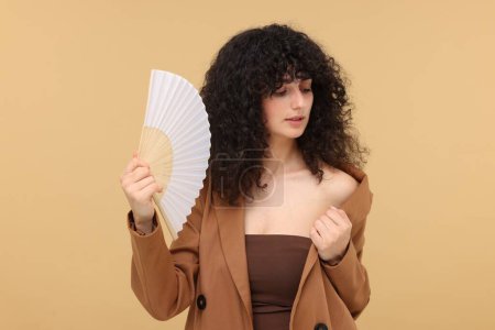 Woman with hand fan suffering from heat on beige background