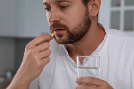 Depressed man with glass of water taking antidepressant pill indoors, closeup