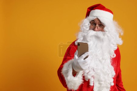 Frohe Weihnachten. Weihnachtsmann mit Smartphone auf orangefarbenem Hintergrund, Platz für Text