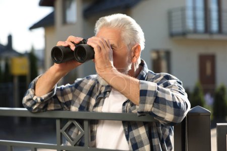 Konzept des Privatlebens. Neugieriger Senior mit Fernglas spioniert Nachbarn über Zaun im Freien aus