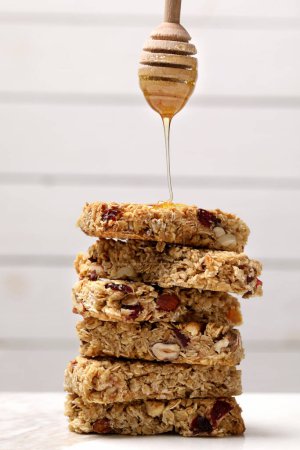 Téléchargez les photos : Verser le miel de la trempette sur une pile de barres de granola savoureuses sur une table blanche - en image libre de droit