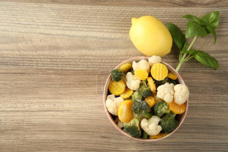 Frozen vegetables in bowl, basil and lemon on wooden table, flat lay. Space for text