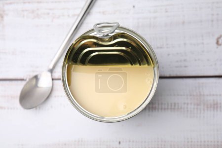 Tasty condensed milk in tin can and spoon on white wooden table, top view