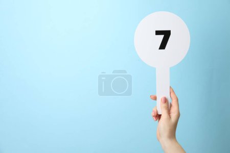 Woman holding auction paddle with number 7 on light blue background, closeup. Space for text