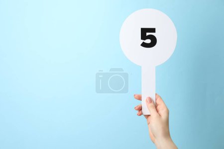 Woman holding auction paddle with number 5 on light blue background, closeup. Space for text