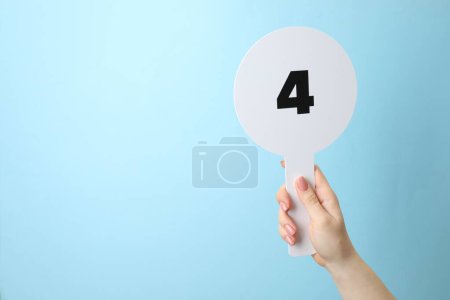 Woman holding auction paddle with number 4 on light blue background, closeup. Space for text