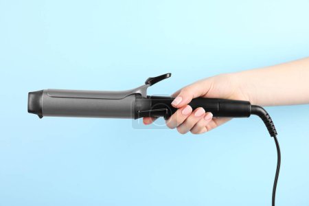 Hair styling appliance. Woman holding curling iron on light blue background, closeup