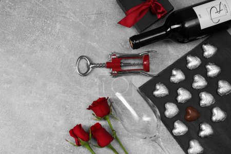 Bottle of red wine, glass, heart shaped chocolate candies, corkscrew, roses and gift box on light grey textured table, flat lay. Space for text