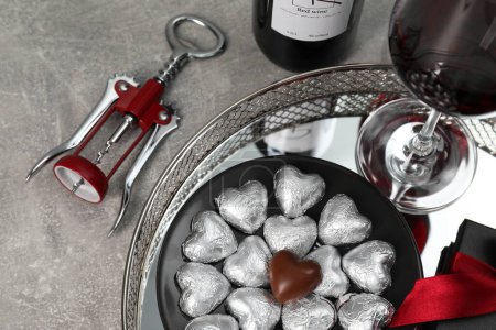 Flasche, Glas Rotwein, herzförmige Schokoladenbonbons, Korkenzieher und Geschenkkarton auf hellgrau strukturiertem Tisch, oben Ansicht