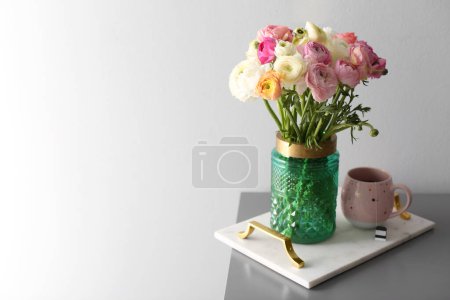 Strauß schöner Hahnenfußgewächse und Tee auf dem Tisch neben hellgrauer Wand. Raum für Text