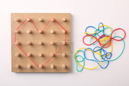 Wooden geoboard with heart made of rubber bands on white table, flat lay. Educational toy for motor skills development